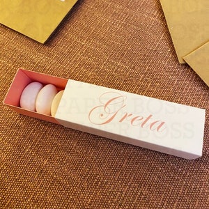 Op de afbeelding: Een witte doos met een roze deksel en de naam "Greta" in roze schrift. De doos bevat vier roze en witte amandelvormige snoepjes.