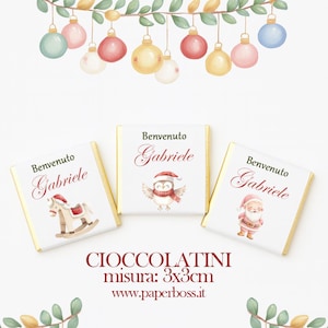 Puede incluir: Tres pequeñas barras de chocolate con bordes de papel dorado y envoltorios blancos. Cada envoltorio presenta una ilustración navideña y el texto "Benvenuto Gabriele". Las barras miden aproximadamente 3 x 3 cm.