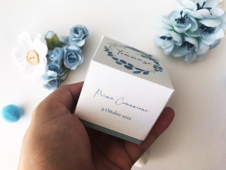 May include: A white gift box with a blue and white floral design. The box has the text "Tommaso" and "Prima Comunione 9 Ottobre 2022" printed on it.