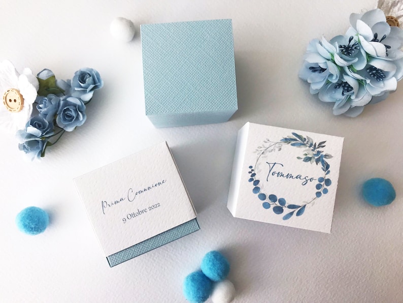 May include: Two white gift boxes with blue accents. One box has the text "Prima Comunione 9 Ottobre 2022" and the other box has the text "Tommaso" surrounded by a blue floral wreath.