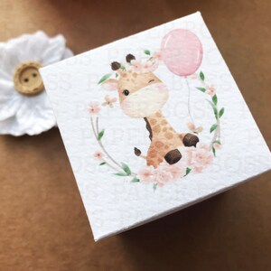 Peut inclure: Une bo&icirc;te carr&eacute;e blanche avec une illustration aquarelle d'une girafe portant une couronne de fleurs et tenant un ballon rose. La girafe est entour&eacute;e de fleurs roses et de feuilles vertes.