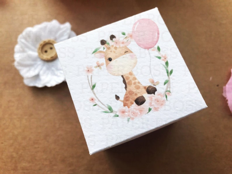 Peut inclure: Une bo&icirc;te carr&eacute;e blanche avec une illustration aquarelle d'une girafe portant une couronne de fleurs et tenant un ballon rose. La girafe est entour&eacute;e de fleurs roses et de feuilles vertes.