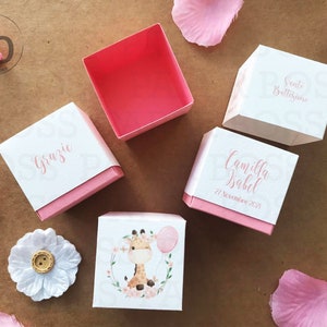Peut inclure: Bo&icirc;tes de drag&eacute;es roses et blanches avec un motif de girafe. Les bo&icirc;tes portent les inscriptions "Grazie", "Camilla Isabel", "21 Novembre 2021" et "Santo Battesimo".