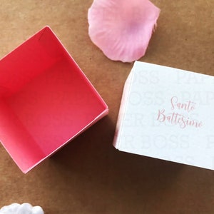 Peut inclure: Une bo&icirc;te cadeau blanche avec les mots "Santo Battesimo" imprim&eacute;s en rose. La bo&icirc;te est ouverte et l'int&eacute;rieur est rose.