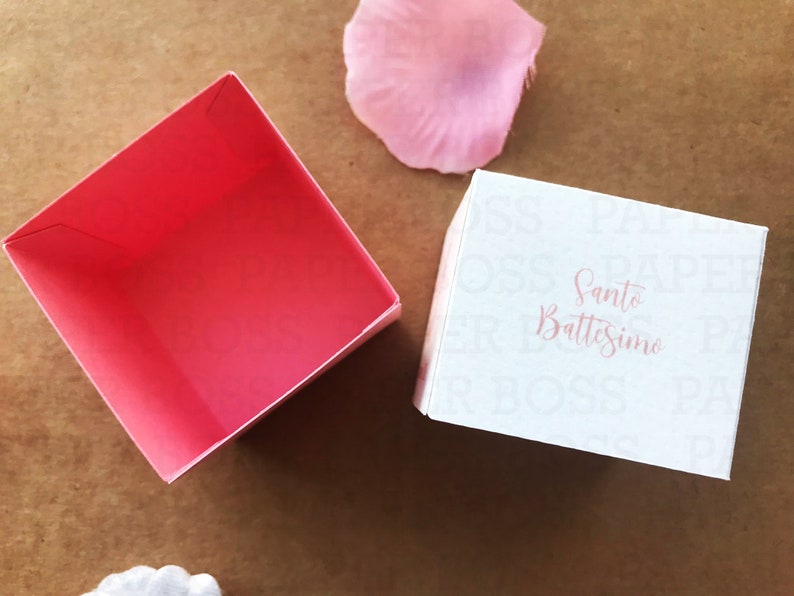 Peut inclure: Une bo&icirc;te cadeau blanche avec les mots "Santo Battesimo" imprim&eacute;s en rose. La bo&icirc;te est ouverte et l'int&eacute;rieur est rose.