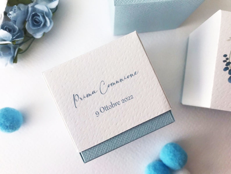 May include: A white gift box with a blue lid. The box is decorated with the text "Prima Comunione" and the date "9 Ottobre 2022".