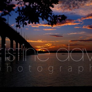 May include: Silhouette of a bridge over water at sunset. The sky is filled with orange, yellow, and blue hues. The water reflects the colors of the sky. The image includes the text "tristine davis photography".