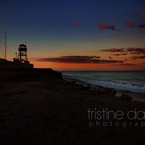 May include: A scenic sunset over a beach with a silhouetted palm tree and a watchtower. The sky transitions from deep blue to orange and red, reflecting on the ocean waves. The beach is dark, with the photographer's name in white.