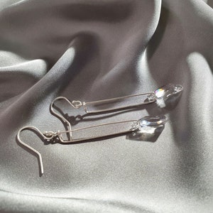 May include: A pair of silver dangle earrings with clear crystal teardrop accents. The earrings feature a simple, straight bar design.