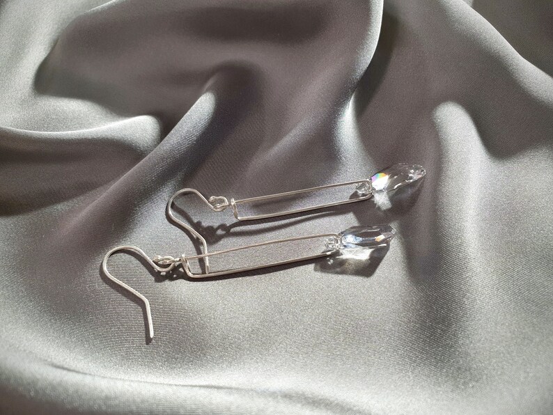 May include: A pair of silver dangle earrings with clear crystal teardrop accents. The earrings feature a simple, straight bar design.
