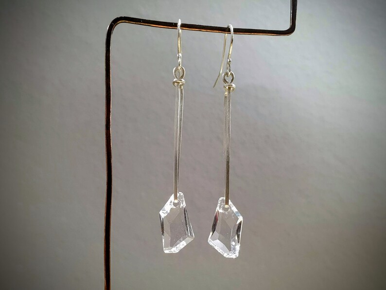 May include: A pair of silver dangle earrings with clear crystal geometric pendants. The earrings are hanging from a silver stand.
