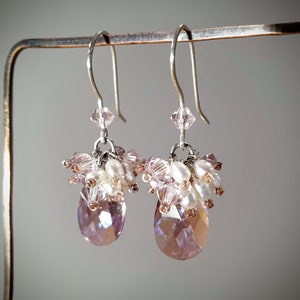May include: A pair of dangle earrings with a teardrop-shaped crystal in shades of pink and purple. The earrings are made with silver-toned metal and feature a cluster of small pink and white beads.