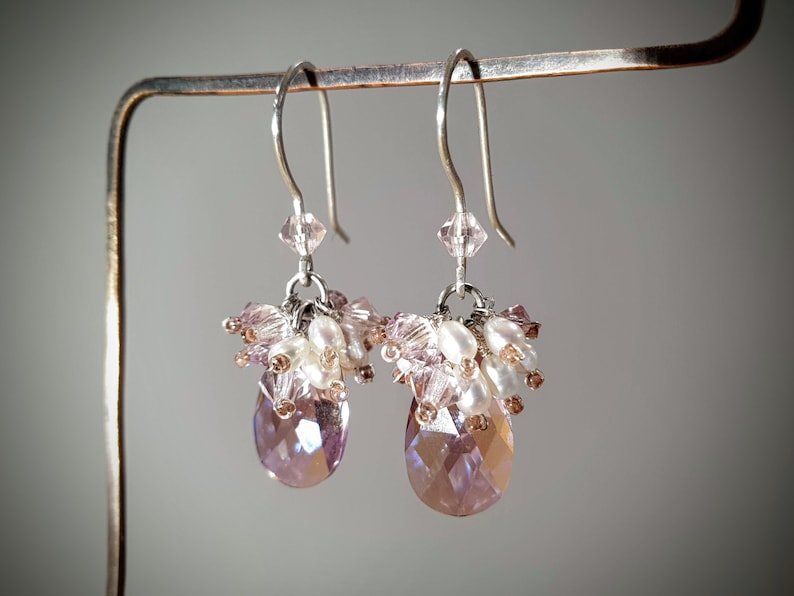 May include: A pair of dangle earrings with a teardrop-shaped crystal in shades of pink and purple. The earrings are made with silver-toned metal and feature a cluster of small pink and white beads.