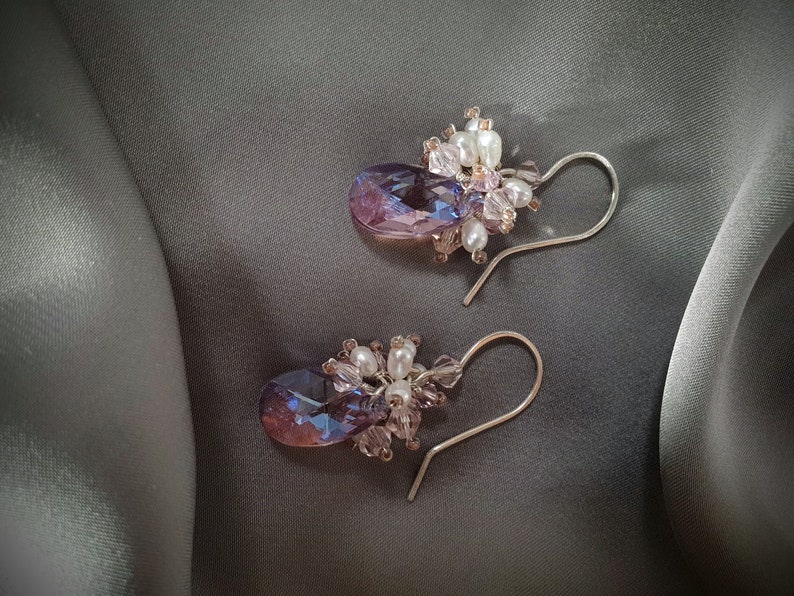 May include: A pair of dangle earrings with a teardrop-shaped purple and blue crystal accented with white pearls and pink beads.