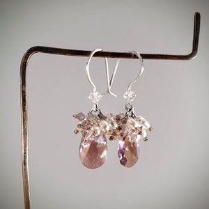 May include: A pair of dangle earrings with a teardrop-shaped crystal in shades of pink and purple. The earrings are adorned with small white pearls and pink and clear crystals.