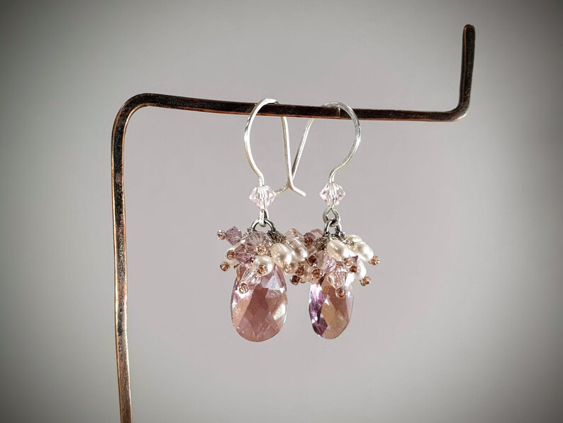 May include: A pair of dangle earrings with a teardrop-shaped crystal in shades of pink and purple. The earrings are adorned with small white pearls and pink and clear crystals.