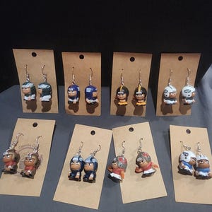 May include: A collection of handmade football player earrings. Each pair features a miniature player figurine in various team colors, attached to silver-tone earring hooks. The earrings are displayed on small, brown cardboard tags.