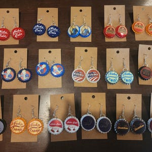 Op de afbeelding: Een display van 10 paar oorbellen gemaakt van flessendoppen. De flessendoppen hebben verschillende ontwerpen, waaronder de woorden "Nuka Cola", "Dark", "Orange", "Victory", "Twist", "Sunset", "Wild", "Cherry" en "Duanitan".