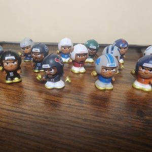 May include: A collection of miniature football player figurines. Each figure is wearing a helmet and uniform representing different NFL teams. The figures are in various poses, with some holding a football. The color scheme includes team colors like blue, red, and white.