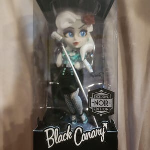 May include: A DC Comics Bombshells Black Canary vinyl figure in a display box. The figure features a character with white hair, a teal and black outfit, and fishnet stockings, holding a microphone. The box includes text: Exclusive Noir Edition.