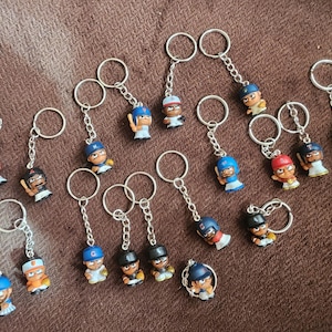 May include: A selection of baseball-themed keyrings, each with a miniature baseball player figurine. The keyrings have silver-coloured rings and chains. The figurines are in various colours, including blue, red, black, and tan.