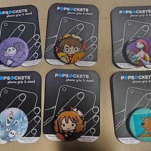 May include: Six different phone grip and stand PopSockets with various cartoon characters. The characters include: Jack Skellington, Harry Potter, Sally, Olaf, Hermione Granger, and Scooby Doo.