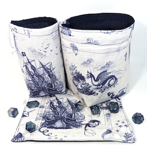 May include: A set of three blue and white fabric dice bags with a nautical theme. The bags feature a ship, an octopus, and a sea serpent. The bags are lined with black fabric and have a drawstring closure. The set also includes a matching dice tray with a nautical design.
