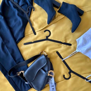 May include: A flat lay of clothing items on a yellow surface. The items include a pair of black pants, a white tank top, a black leather handbag with a gold chain strap, and three black hangers. The handbag has a tag that says "Christian Dior Paris".