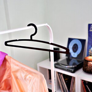 May include: A black plastic clothes hanger hangs on a white clothing rack. A peach-colored jacket with purple accents is partially visible. Shelves in the background hold vinyl records and a record player, suggesting a music or fashion theme.