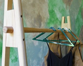 Könnte beinhalten: Ein Kleiderständer aus Holz mit natürlicher Oberfläche. Der Ständer hat eine horizontale Stange und zwei schräge Beine. Drei grüne Plastikbügel hängen an der Stange, an denen eine braune Jacke und ein schwarzes Tanktop hängen.
