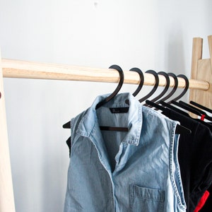 May include: A wooden clothing rack with a natural finish. The rack has a single horizontal bar and two vertical supports. Black hangers with clothing are hanging on the bar.