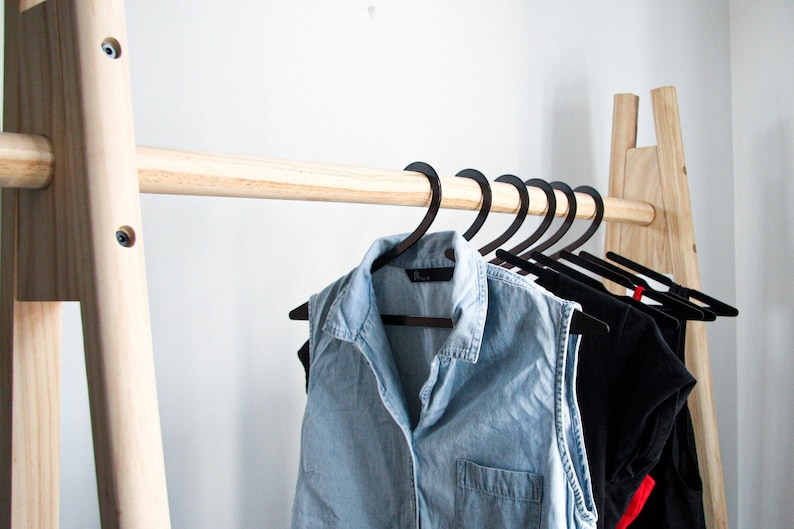 May include: A wooden clothing rack with a natural finish. The rack has a single horizontal bar and two vertical supports. Black hangers with clothing are hanging on the bar.