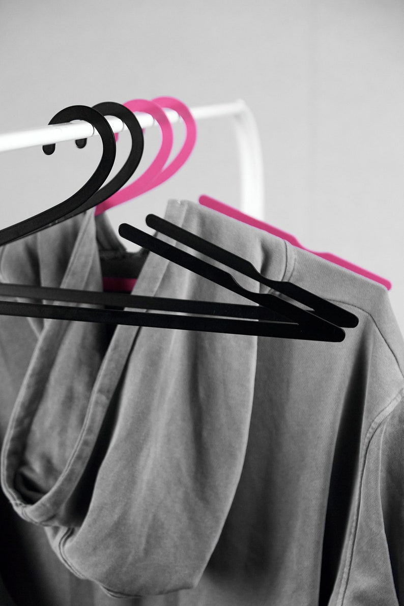 May include: A gray hooded sweatshirt hanging on a white clothes rack with two black and two pink plastic hangers.