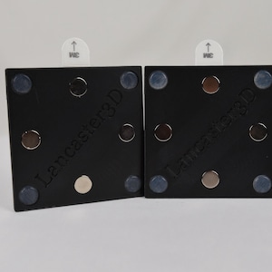 May include: Two square black mounts featuring the text Lancaster3D embossed on the surface. Each mount includes four small circular magnets at the corners, a central circular magnet, and a white adhesive tab with an upward arrow at the top edge.