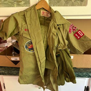 May include: Olive green vintage Boy Scout uniform jacket with patches. Features a red and white patch that says "SHE N.Y. 10". Includes a matching olive green neckerchief. The jacket is hanging on a wooden hanger.