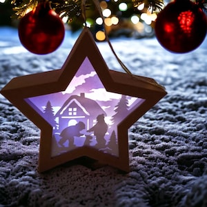 Könnte beinhalten: Ein sternförmiger Weihnachtsschmuck mit einer ausgeschnittenen Winterszene. Der hellbraune Holzschmuck leuchtet innen lila und zeigt eine Silhouette von Haus, Bäumen und Figuren. Rote Kugeln hängen im Hintergrund, auf einem schneebedeckten Boden.
