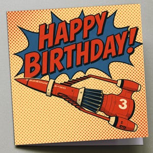 May include: A birthday card in a retro comic book style. The card features a red rocket ship with the number 3 and the words "HAPPY BIRTHDAY!" in bold red letters against a blue burst on a dotted yellow background.