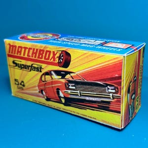 以下が含まれることがあります： ヴィンテージのマッチボックス スーパーファストのおもちゃの車の箱。箱には、赤と黒のフォードカプリ車のイラストが、黄色と赤の背景で描かれています。「MATCHBOX Superfast 54 CAPRI」というテキストが箱に表示されています。