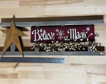 May include: Wooden Christmas decoration with a star and a sign that reads "Believe in Magic". The sign is burgundy with white lettering and snowflake accents. A ruler is placed next to the decoration. The decoration is approximately 24 inches long.