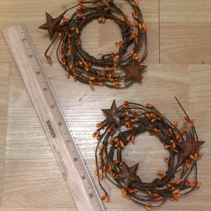 May include: Two rustic, circular wreaths with brown stems, orange berries, and rusty metal star accents. A wooden ruler with inch measurements is next to the wreaths. The wreaths are likely for home decor.