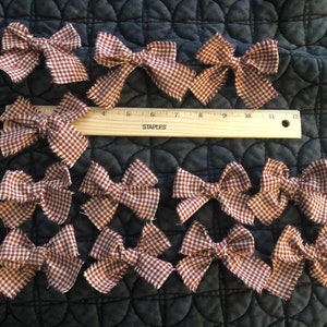 May include: A collection of 12 red and white gingham bows with frayed edges. The bows are arranged in rows on a dark fabric background.