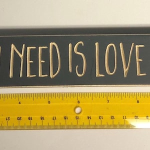 Könnte beinhalten: Ein dunkelgraues Holzschild mit weißer Aufschrift, auf dem "Alles, was du brauchst, ist Liebe und eine Katze" steht.