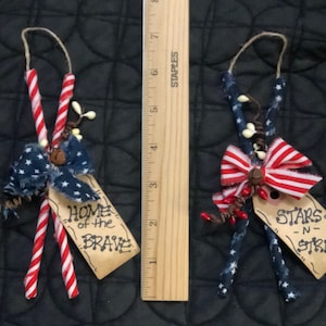 May include: Two patriotic ornaments with red, white, and blue accents. One features a blue bow with white stars and the text "HOME of the BRAVE." The other has a red and white striped bow and reads "STARS -N- STRIPES."