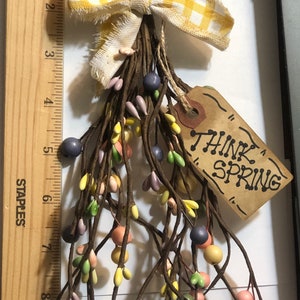 May include: A brown twig spring wreath with yellow, pink, green, and purple berries. A yellow and white gingham bow is tied at the top. A brown tag with the text "Think Spring" hangs from the wreath.