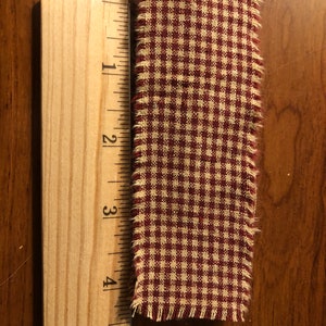 K&ouml;nnte beinhalten: Ein rotes und wei&szlig;es Gingham-Stoffband mit ausgefransten Kanten. Das Band ist etwa 10 cm lang.