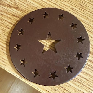 May include: A circular brown metal coaster featuring a large star cutout in the center, surrounded by a ring of twelve smaller star-shaped cutouts arranged evenly around the perimeter of the disc, creating a decorative pattern on the flat surface.