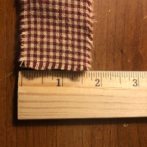 K&ouml;nnte beinhalten: Nahaufnahme eines roten und wei&szlig;en Gingham-Stoffes mit ausgefransten Kanten. Der Stoff wird mit einem Holzlineal gemessen und zeigt, dass er etwa 2,5 cm breit ist.