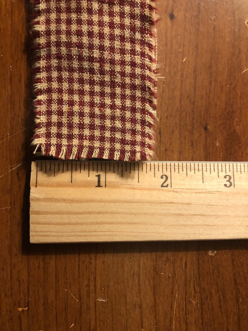 K&ouml;nnte beinhalten: Nahaufnahme eines roten und wei&szlig;en Gingham-Stoffes mit ausgefransten Kanten. Der Stoff wird mit einem Holzlineal gemessen und zeigt, dass er etwa 2,5 cm breit ist.