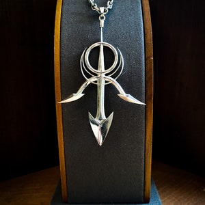 May include: A silver pendant necklace with a geometric design featuring a circle, a cross, and two arrow-like shapes. The pendant is hanging from a silver chain on a black display stand.