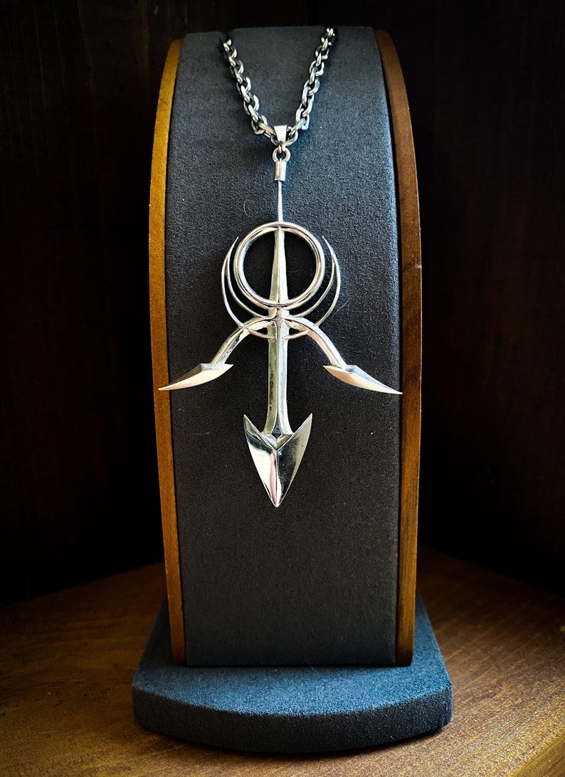 May include: A silver pendant necklace with a geometric design featuring a circle, a cross, and two arrow-like shapes. The pendant is hanging from a silver chain on a black display stand.